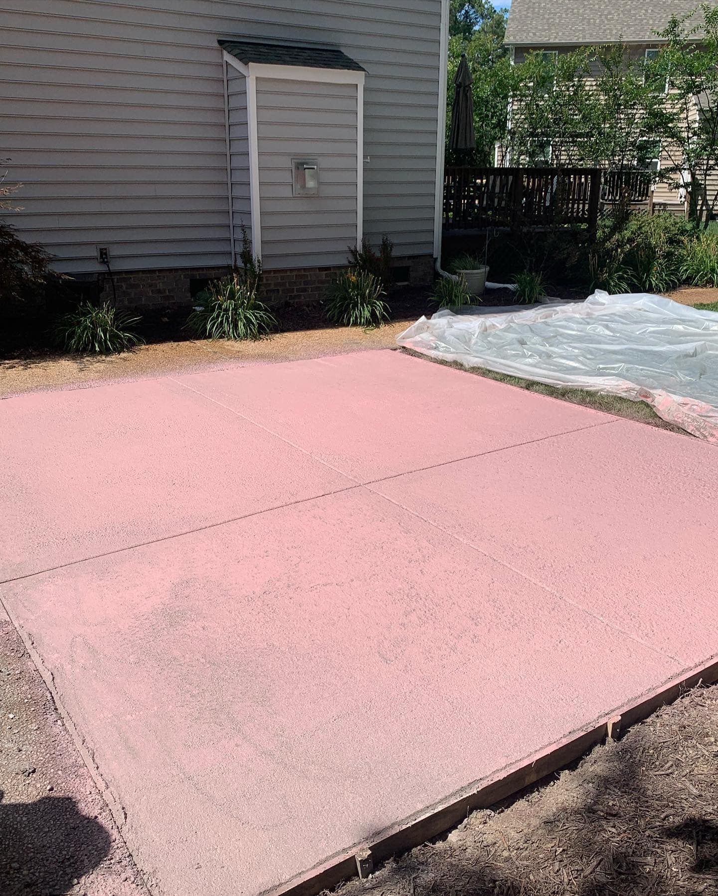 Pink-colored concrete patio with dark border in front of a gray house and small garden.