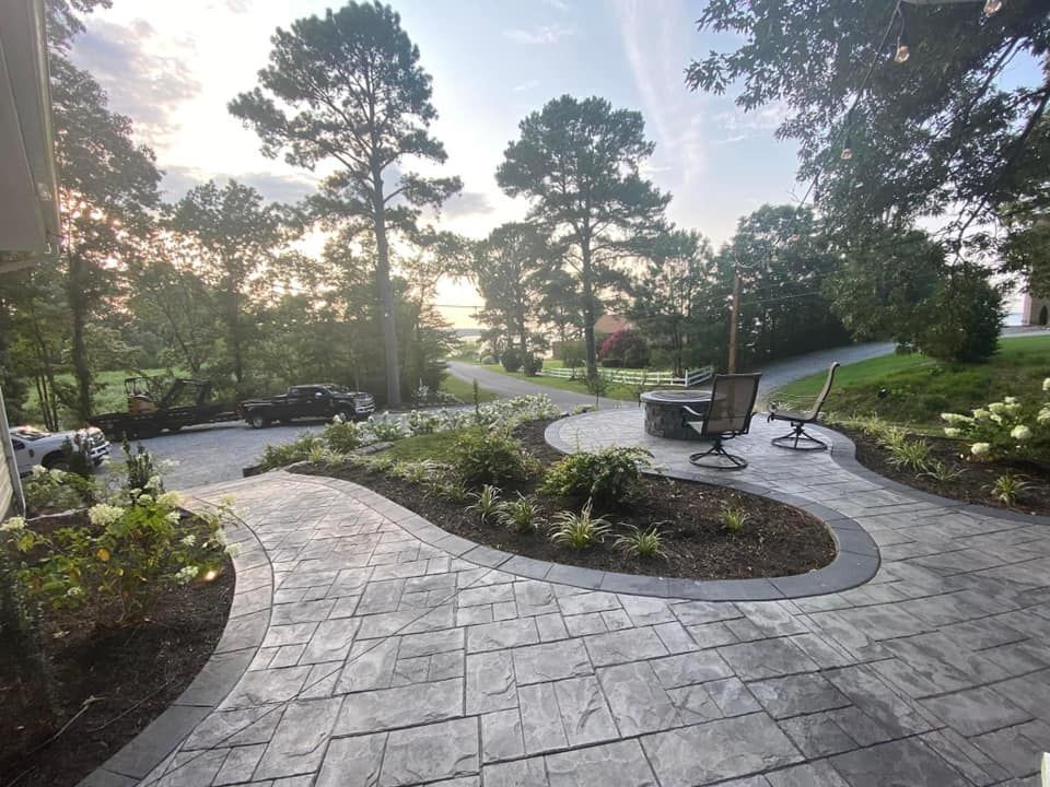 Curved paved patio with fire pit, surrounded by landscaping and trees under a cloudy sky.