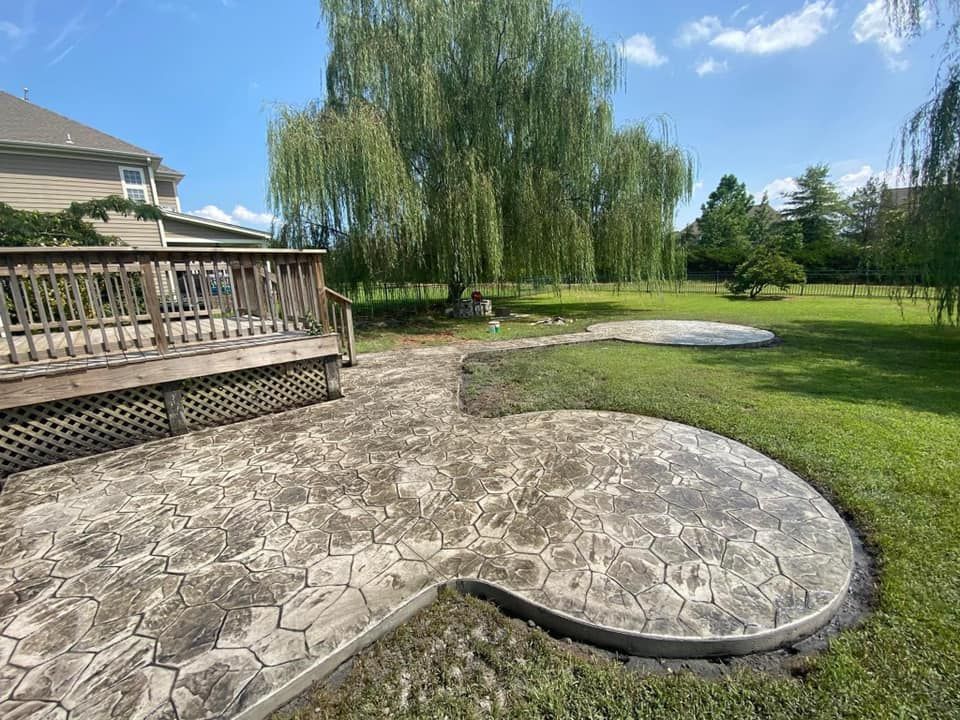 Backyard patio with stamped concrete, deck, large tree, and green lawn on a sunny day.