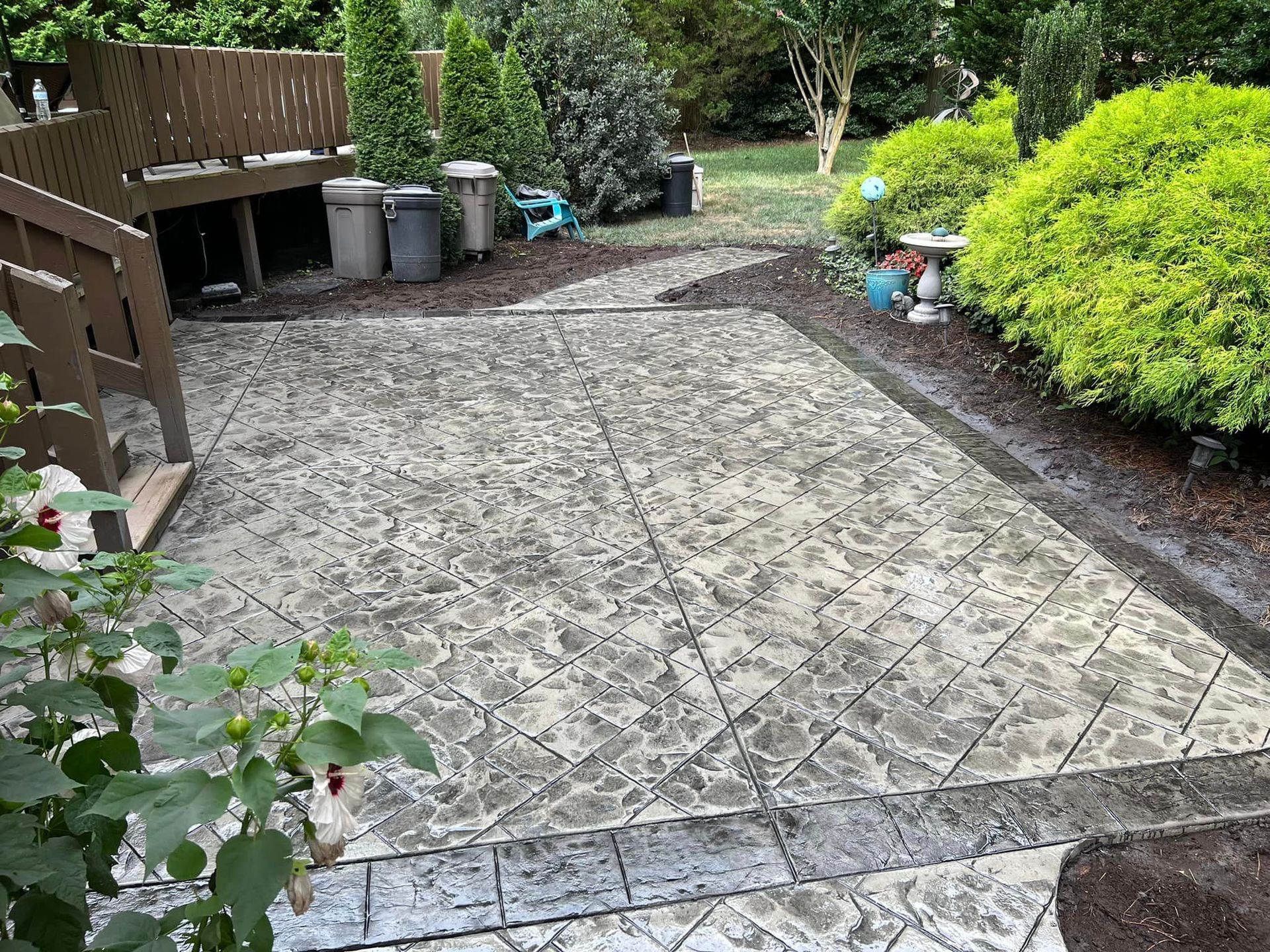 Concrete patio with textured surface, bordering lush greenery and a wooden deck.