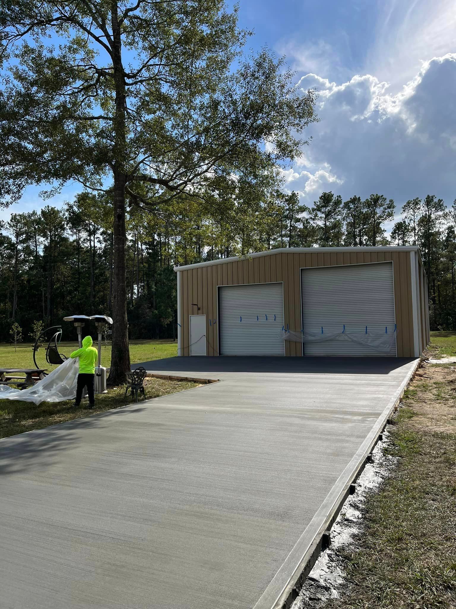 A concrete driveway leads to a tan metal building with two garage doors. A person in a yellow vest stands near a tree.