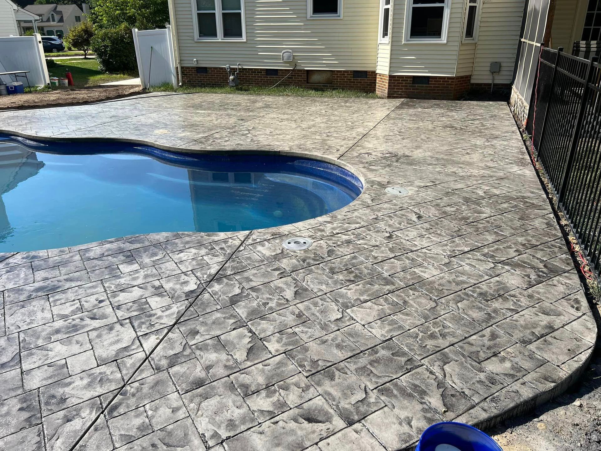 Poolside view of a pool with gray stamped concrete patio. House and black fence visible.