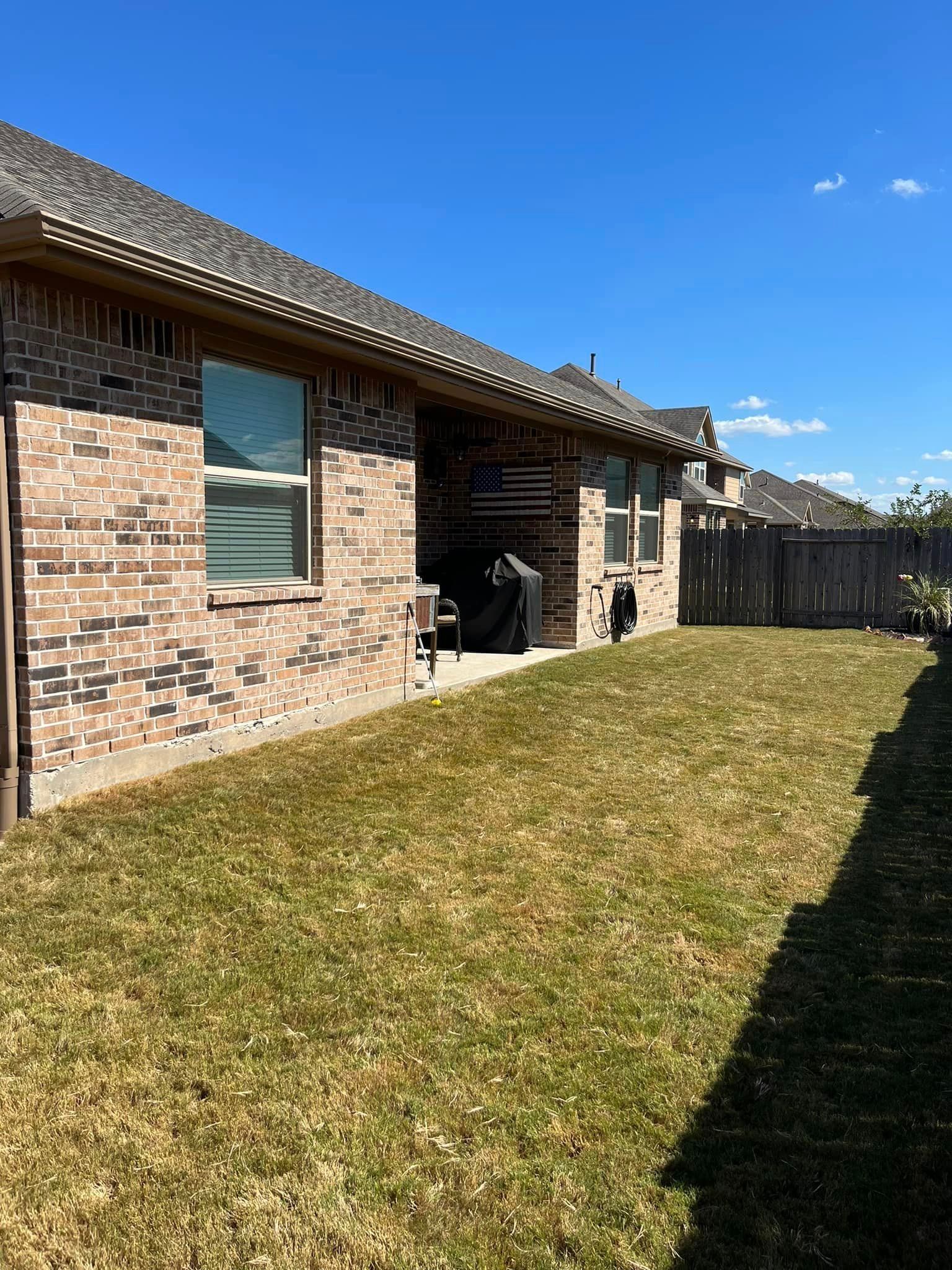 Backyard view: brick house with patio, green grass, blue sky, and wooden fence.