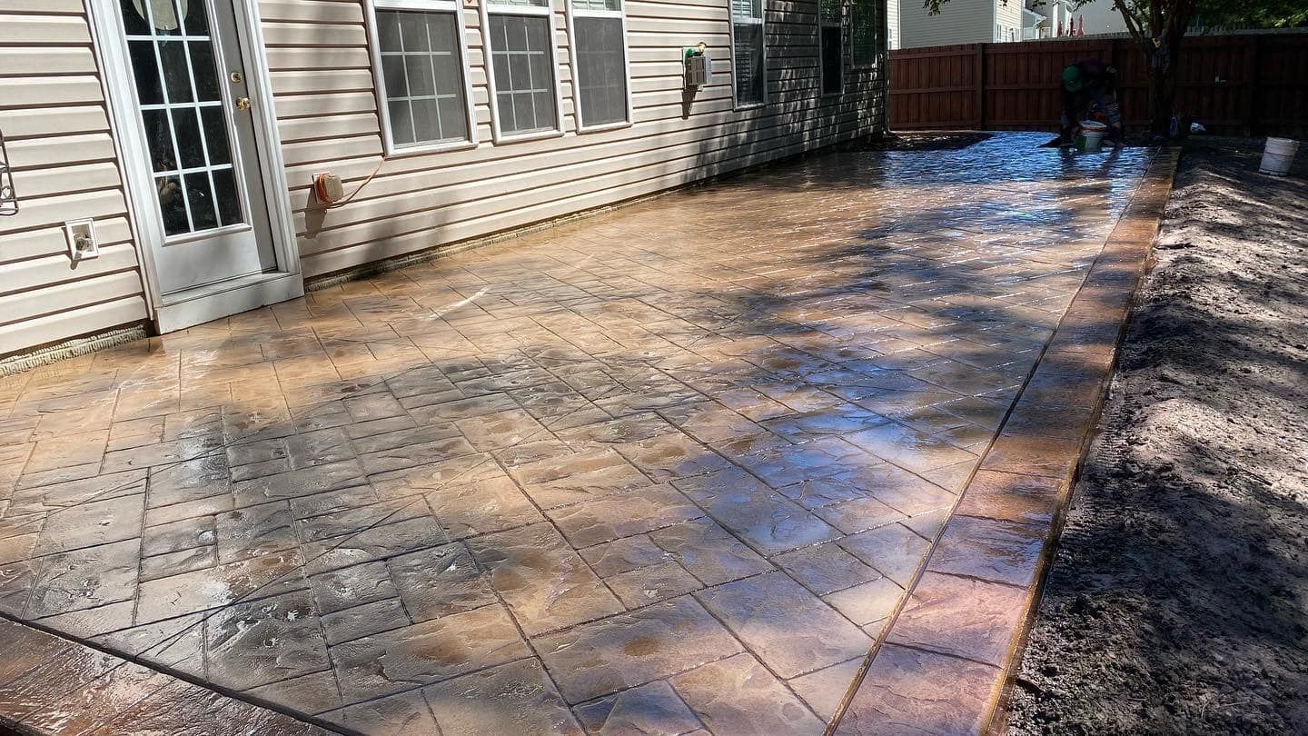 Wet, patterned concrete patio next to a house, edged by a brick border, and surrounded by dirt.