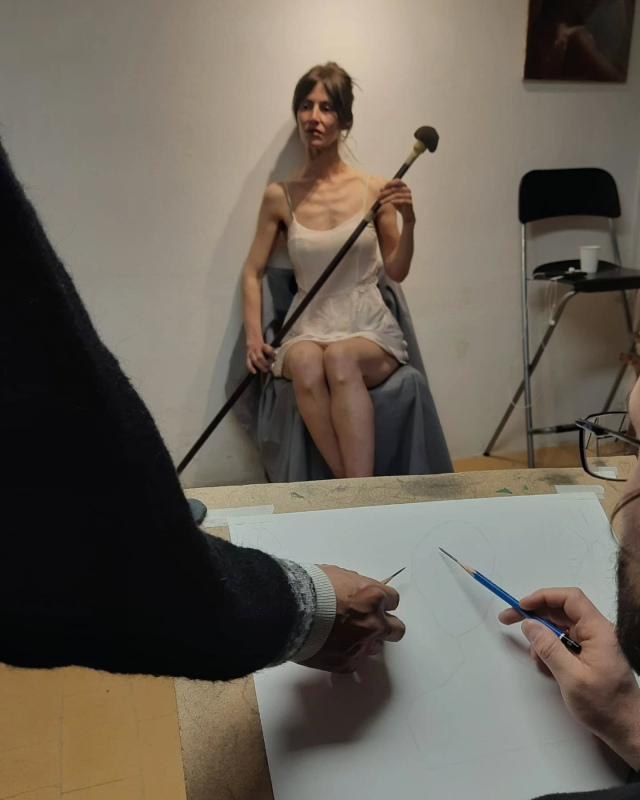 Una persona posa con un bastone per gli artisti. La modella è seduta su una sedia. Due artisti disegnano.