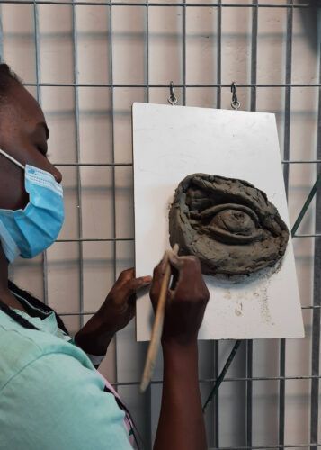 Persona che scolpisce un occhio in argilla su una tavola, indossando una maschera, in un laboratorio.