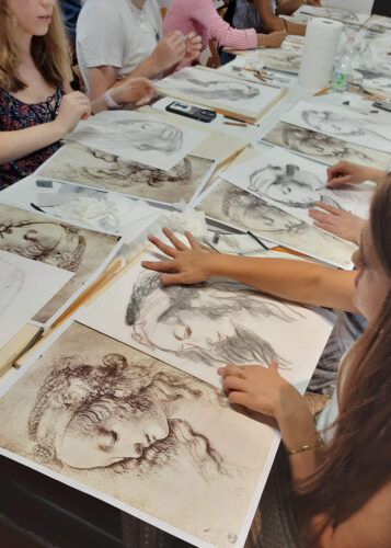 Persone che disegnano a un tavolo; disegni di volti e figure; mani che lavorano; l'ambientazione sembra quella di una lezione o di un laboratorio.