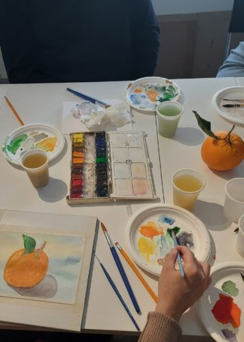 Lezione di pittura ad acquerello: primo piano di una natura morta con frutta arancione, tavolozze, pennelli e bicchieri di vernice.