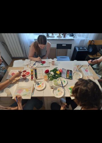 Persone che dipingono seduti a un tavolo con acquerelli, fiori e tavolozze in una stanza ben illuminata.