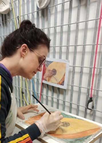Una donna dipinge un liuto, usando una foto di riferimento. Indossa occhiali, guanti e un grembiule da lavoro.