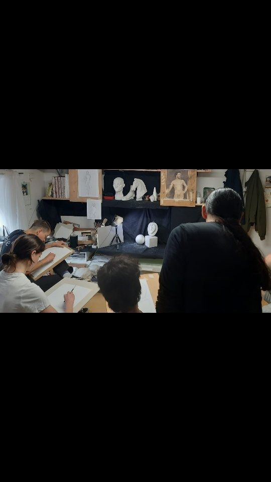 Lezione d'arte: gli studenti disegnano, un insegnante assiste. Luce dalla finestra, modelli sul tavolo.