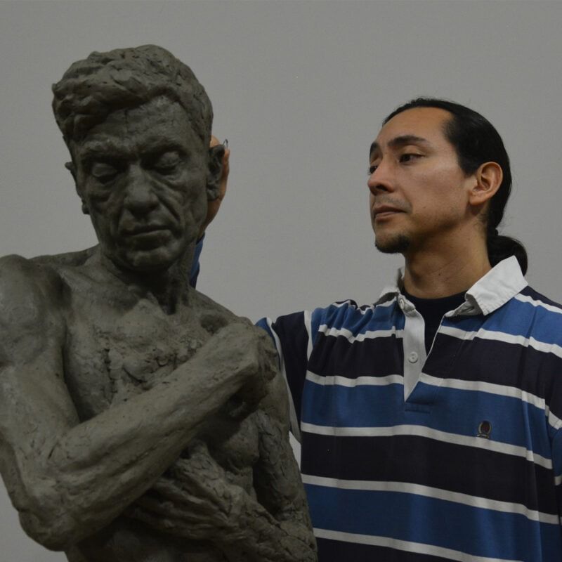 Un uomo con una camicia a righe accanto a una scultura in argilla raffigurante un uomo. Entrambi sono rivolti a sinistra, l'artista tocca la spalla della scultura.