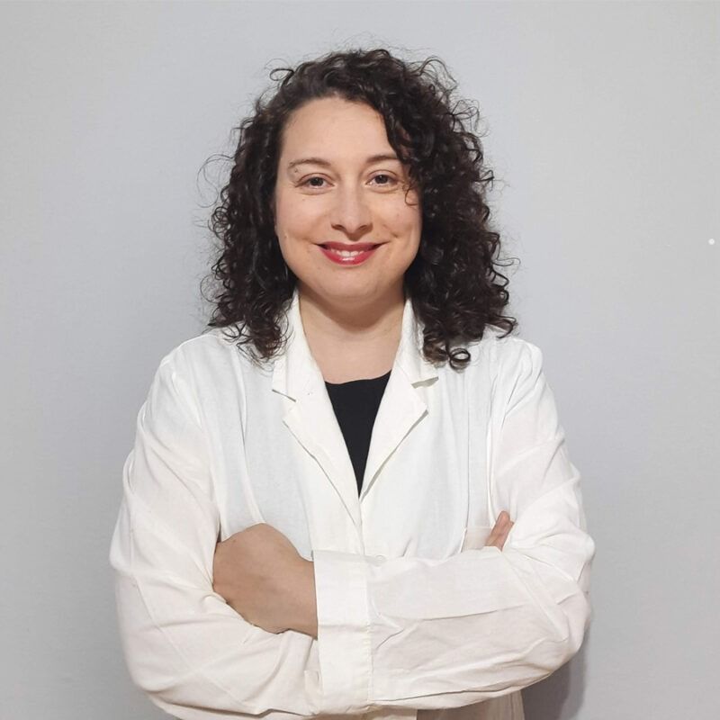 Donna sorridente con capelli castani ricci e braccia incrociate, che indossa un camice bianco.