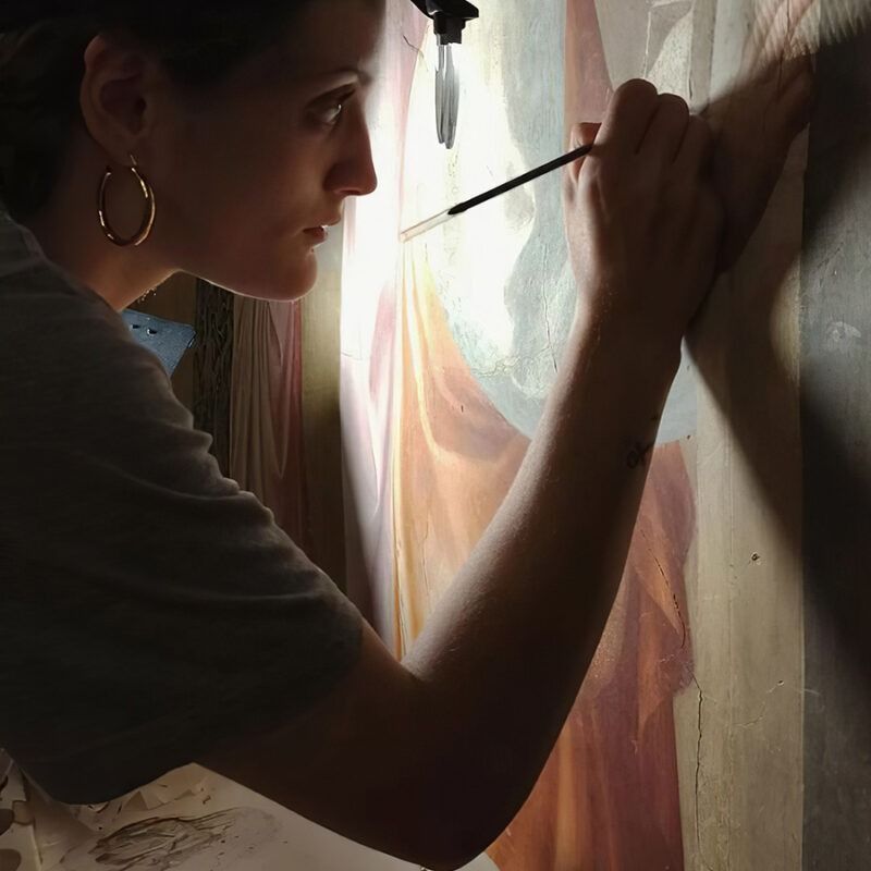 Una donna dipinge un murale, illuminata da una luce zenitale. Indossa orecchini a cerchio d'oro.