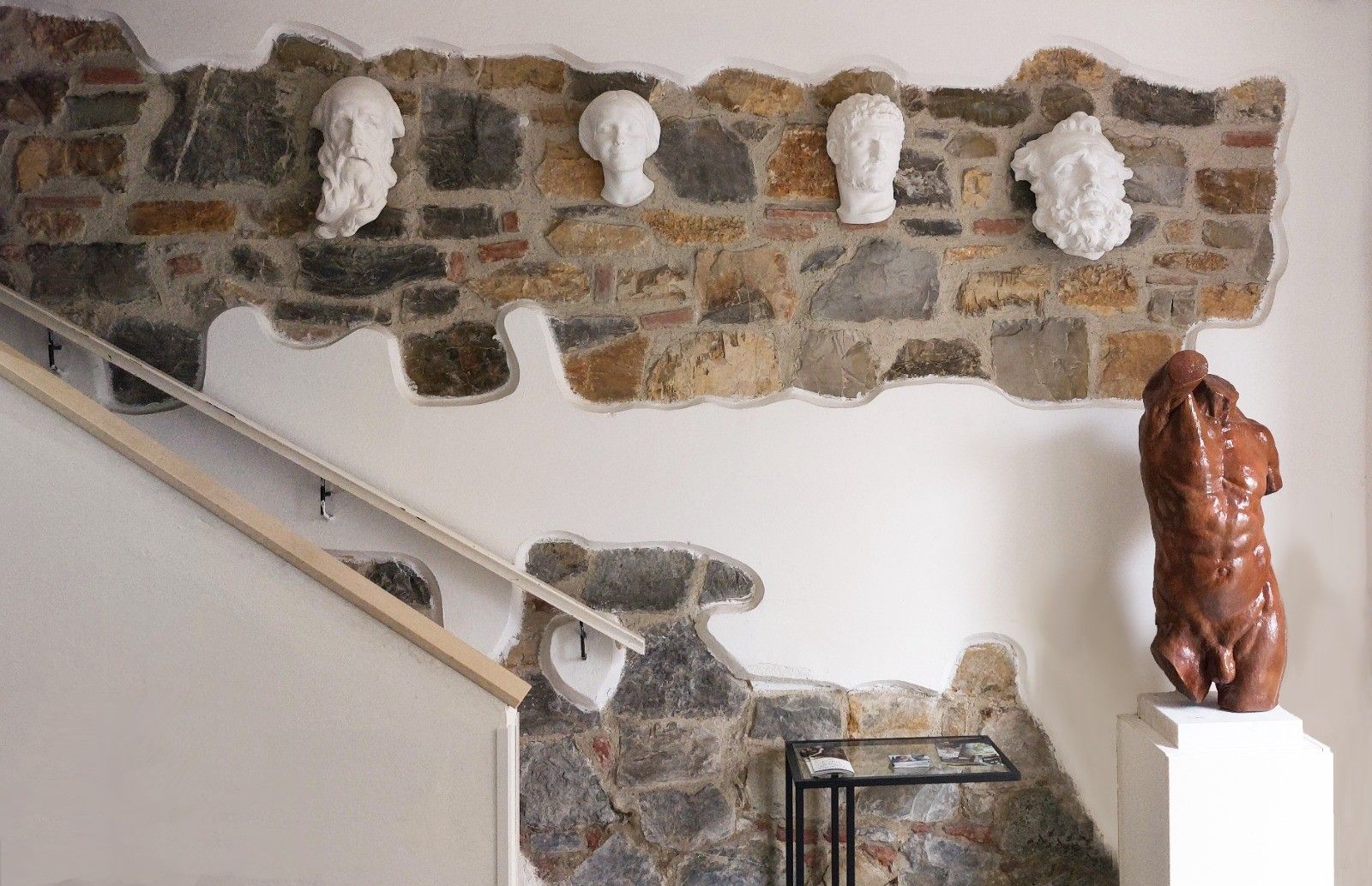 Muro in pietra con ritagli in gesso e busti bianchi, scala e scultura a forma di torso marrone.