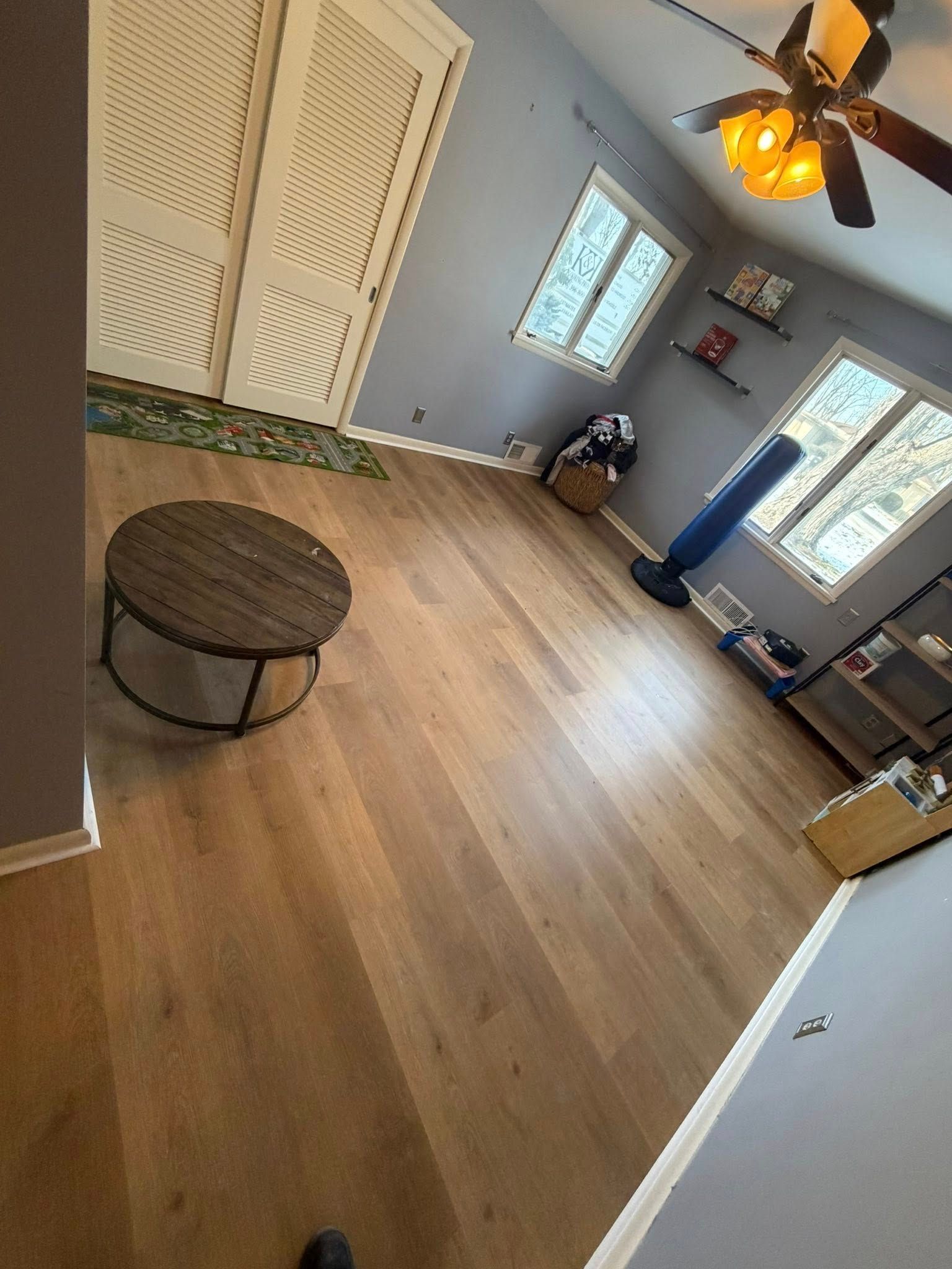 Empty living room with wood floor, ceiling fan, windows, and a small round table.