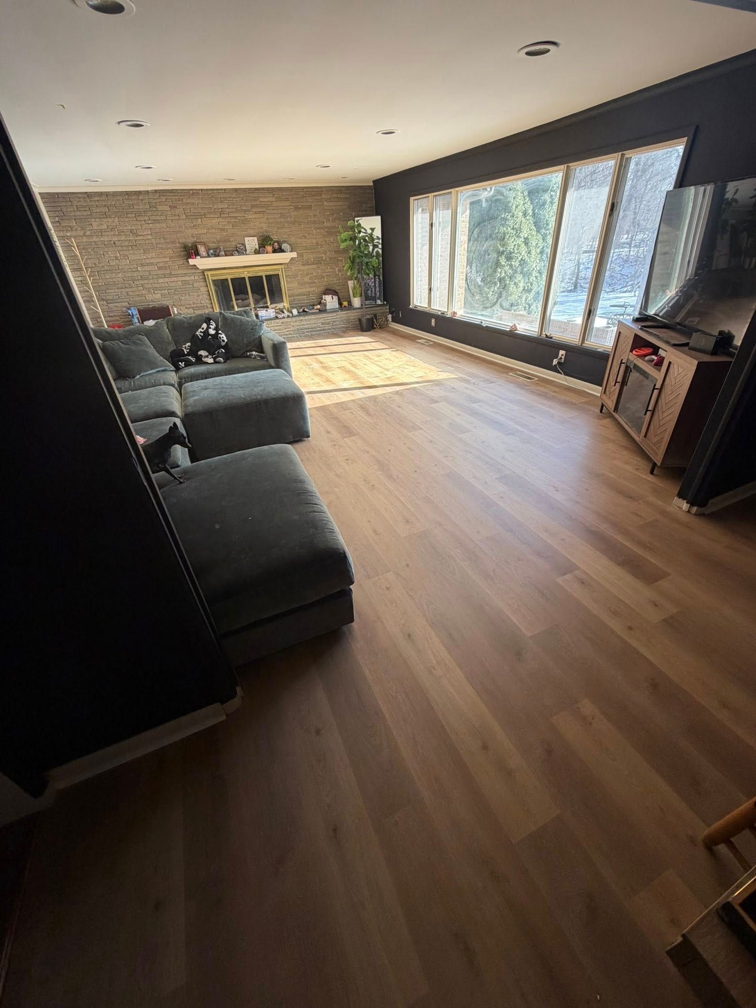 Bright living room with gray sofa, wooden floors, and large windows overlooking trees.