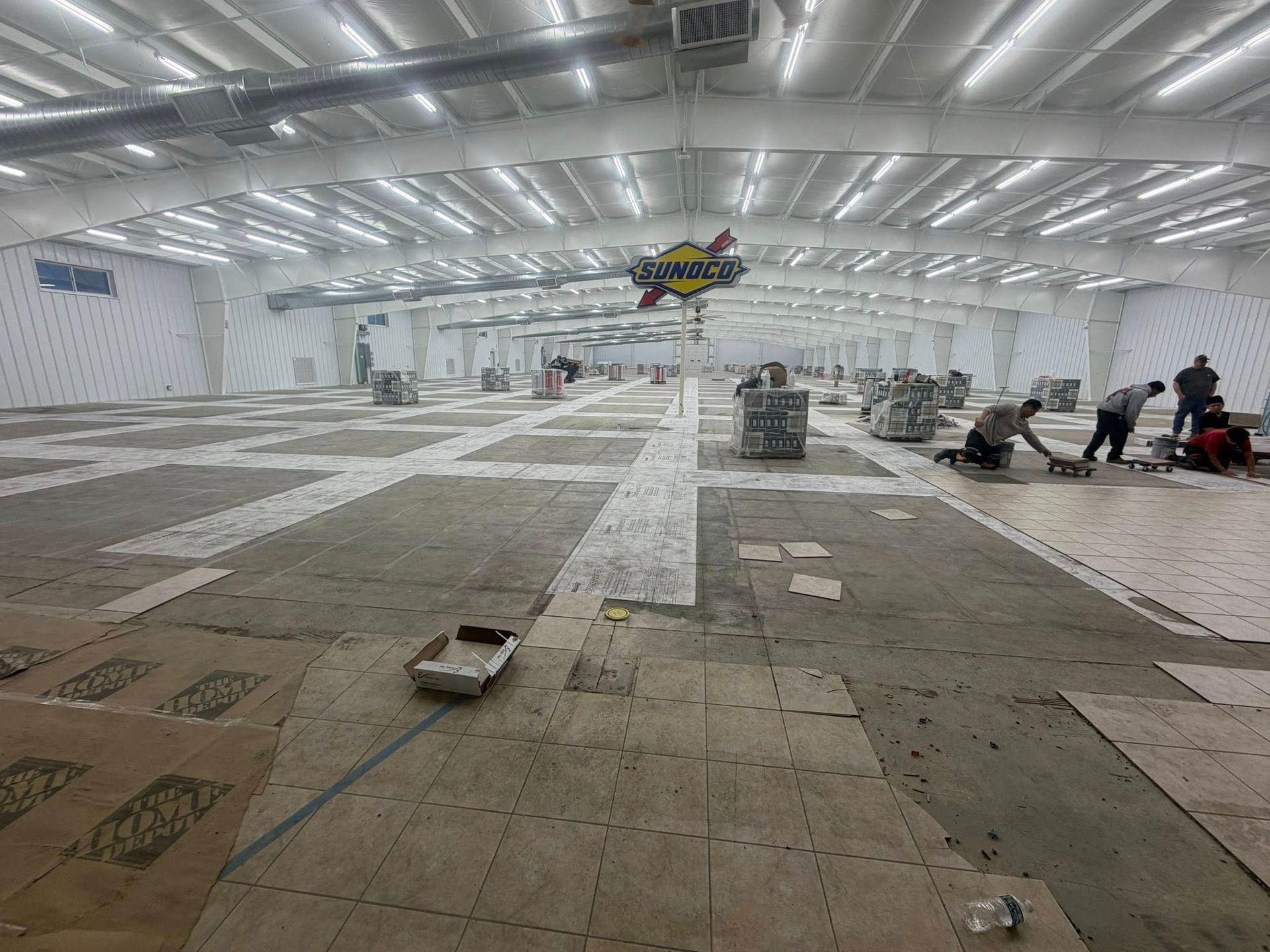 Large indoor hall under construction with workers, white ceiling lights, and materials spread across the floor
