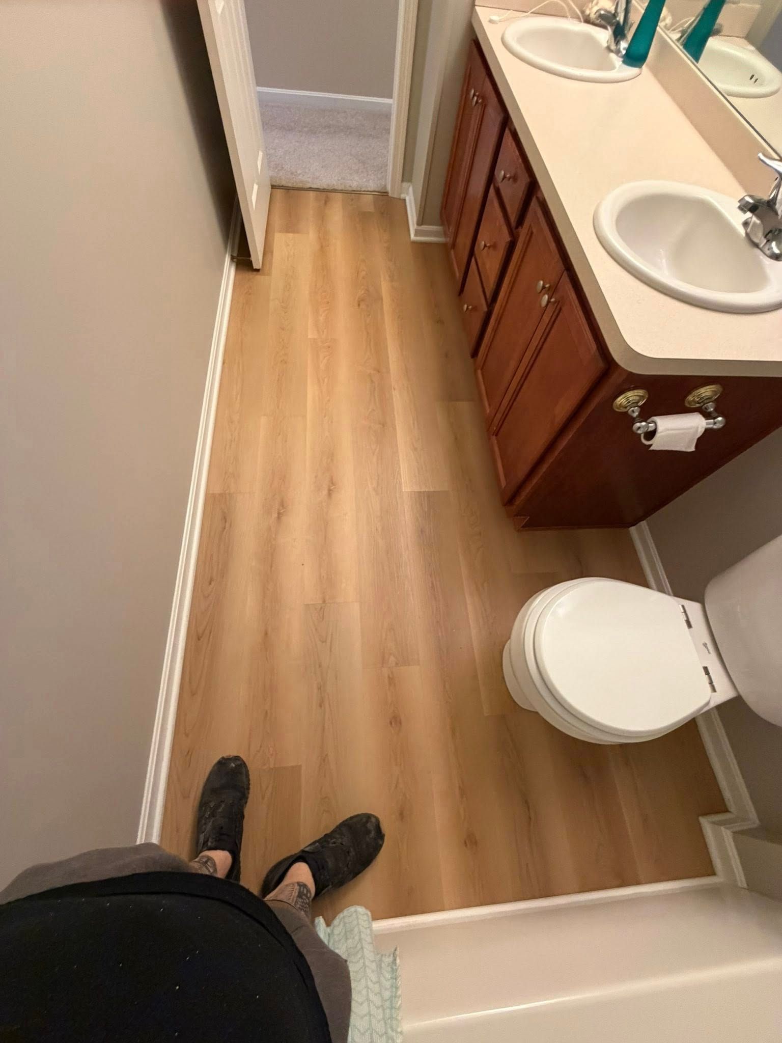 Small bathroom with wood floor, white toilet, double sink vanity, and a person standing in the doorway.