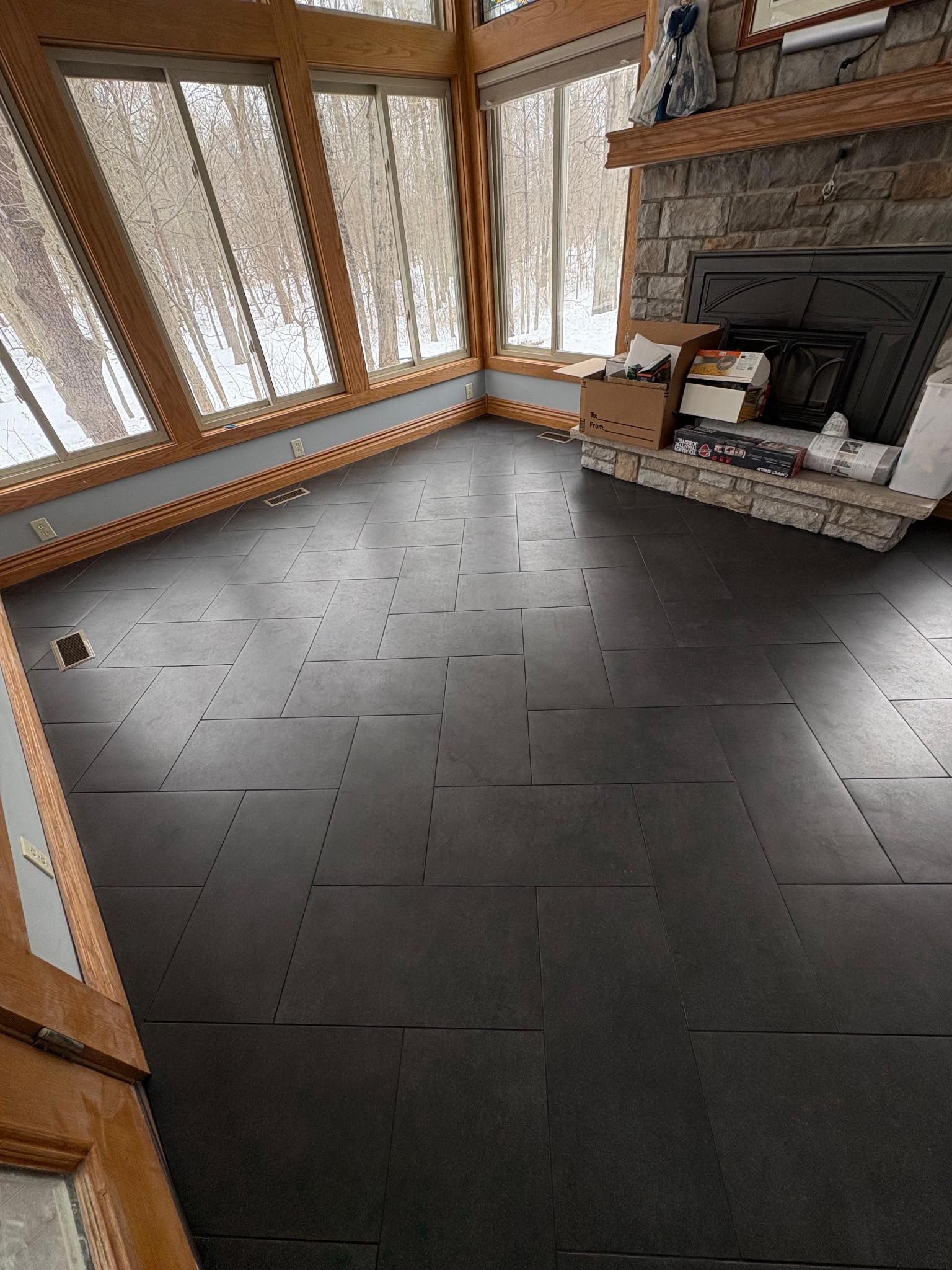 Empty living room with dark tile floor, wood-trimmed windows, and a stone fireplace