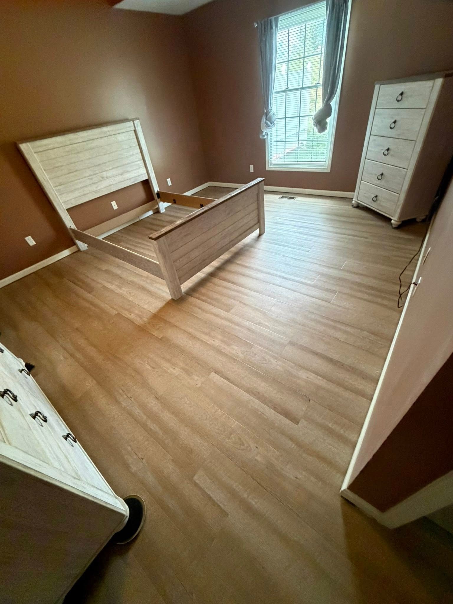 Empty bedroom with brown walls, wood floor, bed frame, window, and dresser.
