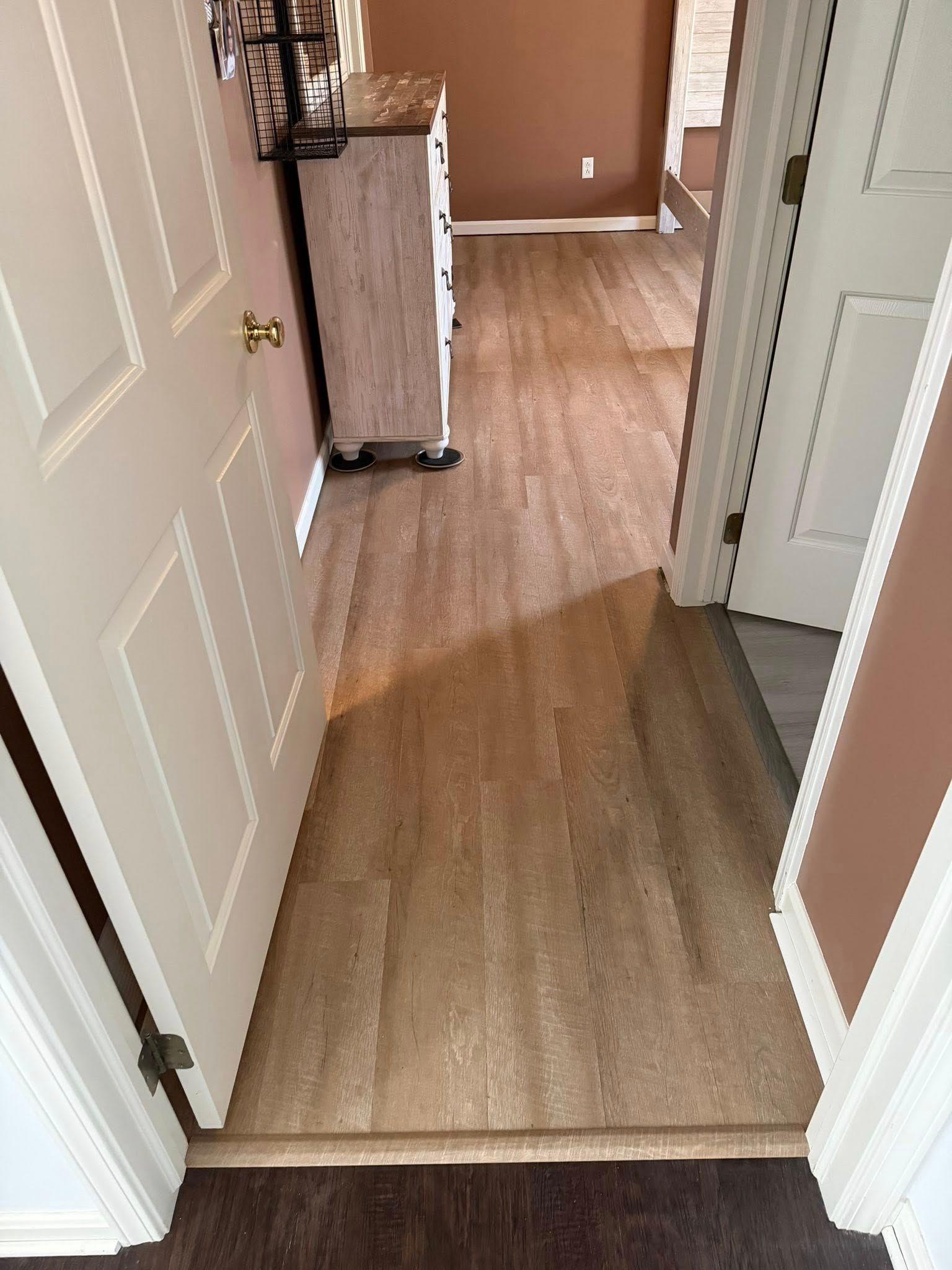 Narrow hallway with light wood flooring and white doors, viewed through an open doorway.