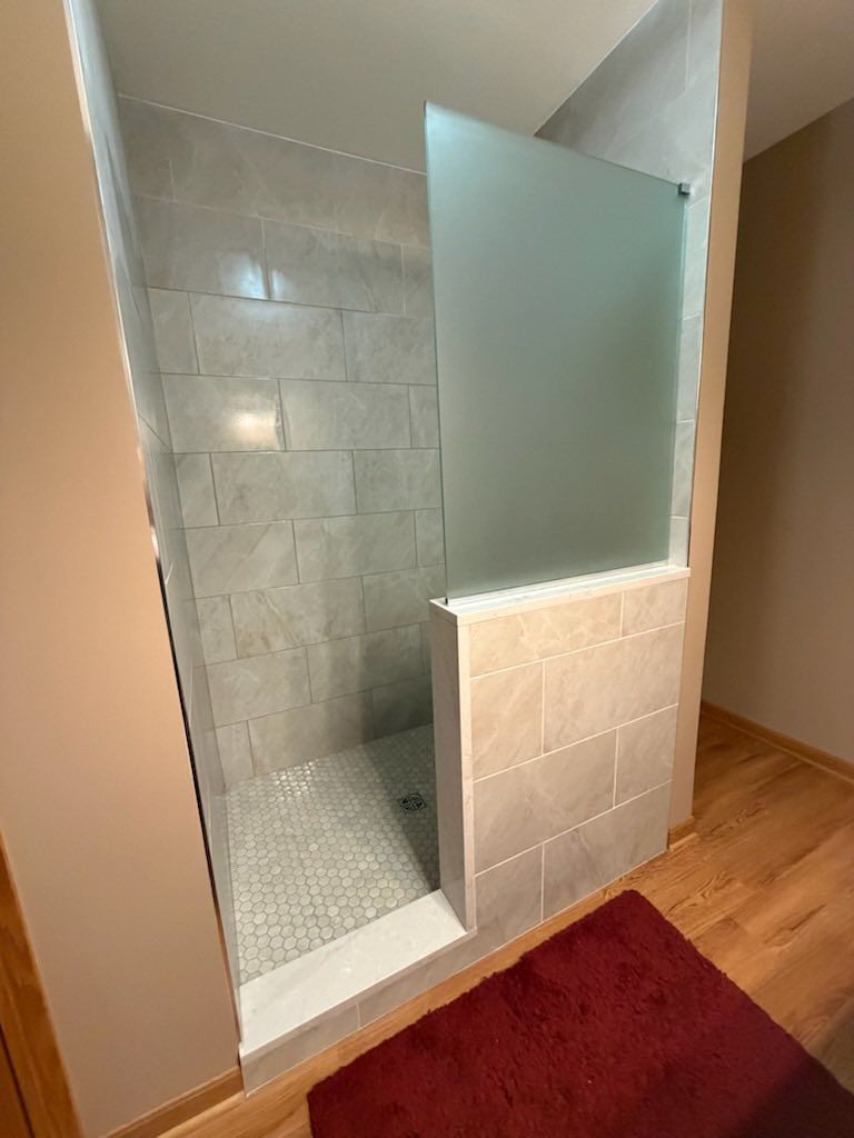 Tiled walk-in shower with glass partition beside a red bath mat in a bathroom