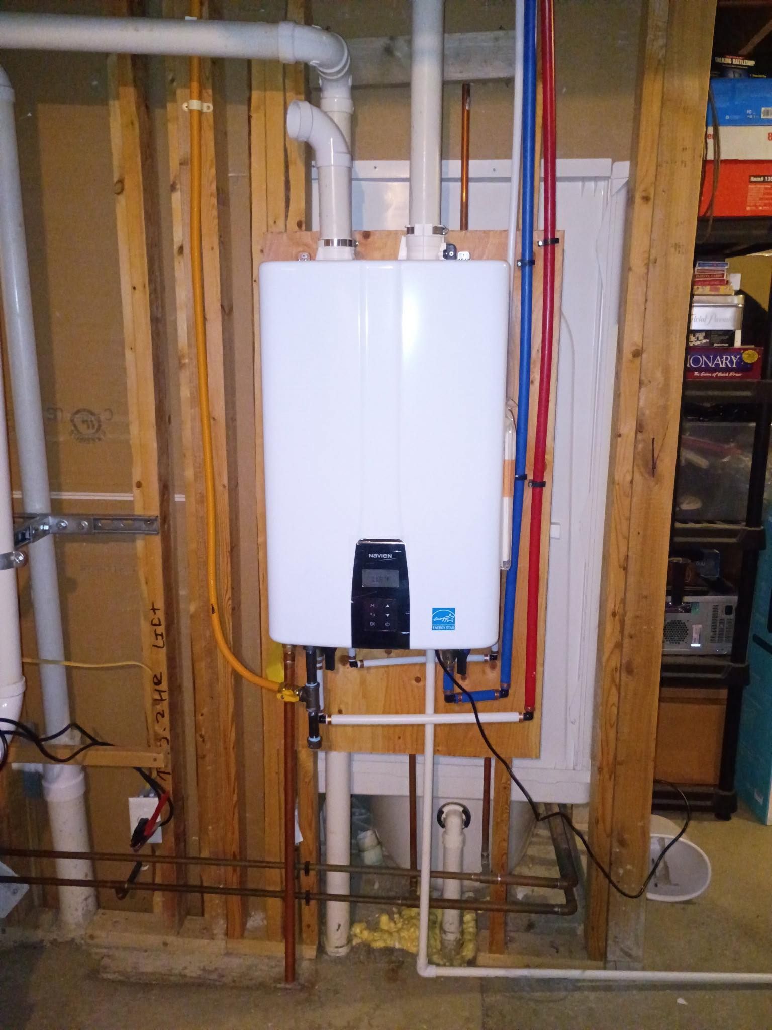 White tankless water heater mounted on exposed basement wall with visible pipes and wiring.