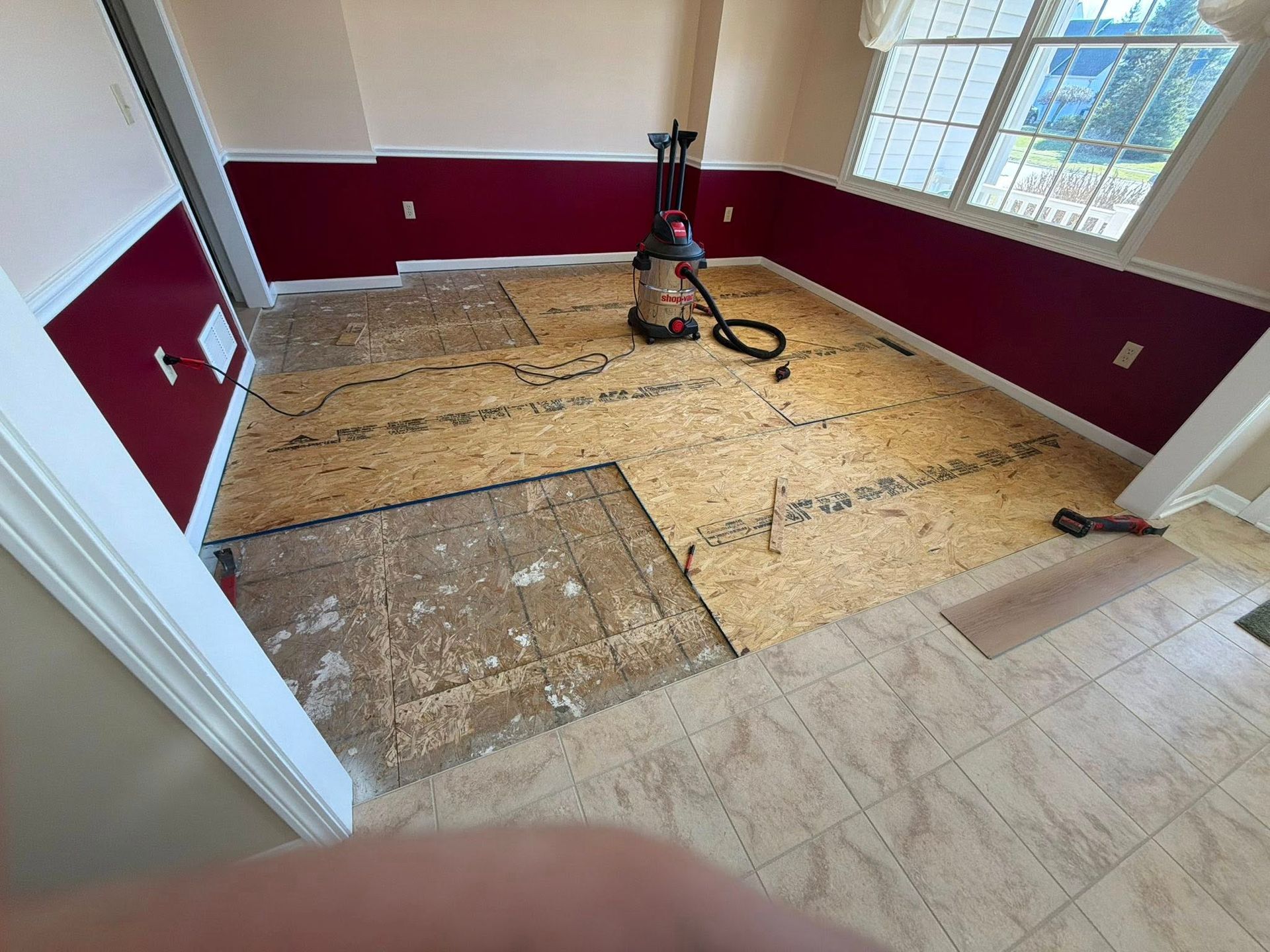 Room under renovation with carpet removed, exposing subfloor and a floor tool near the window.