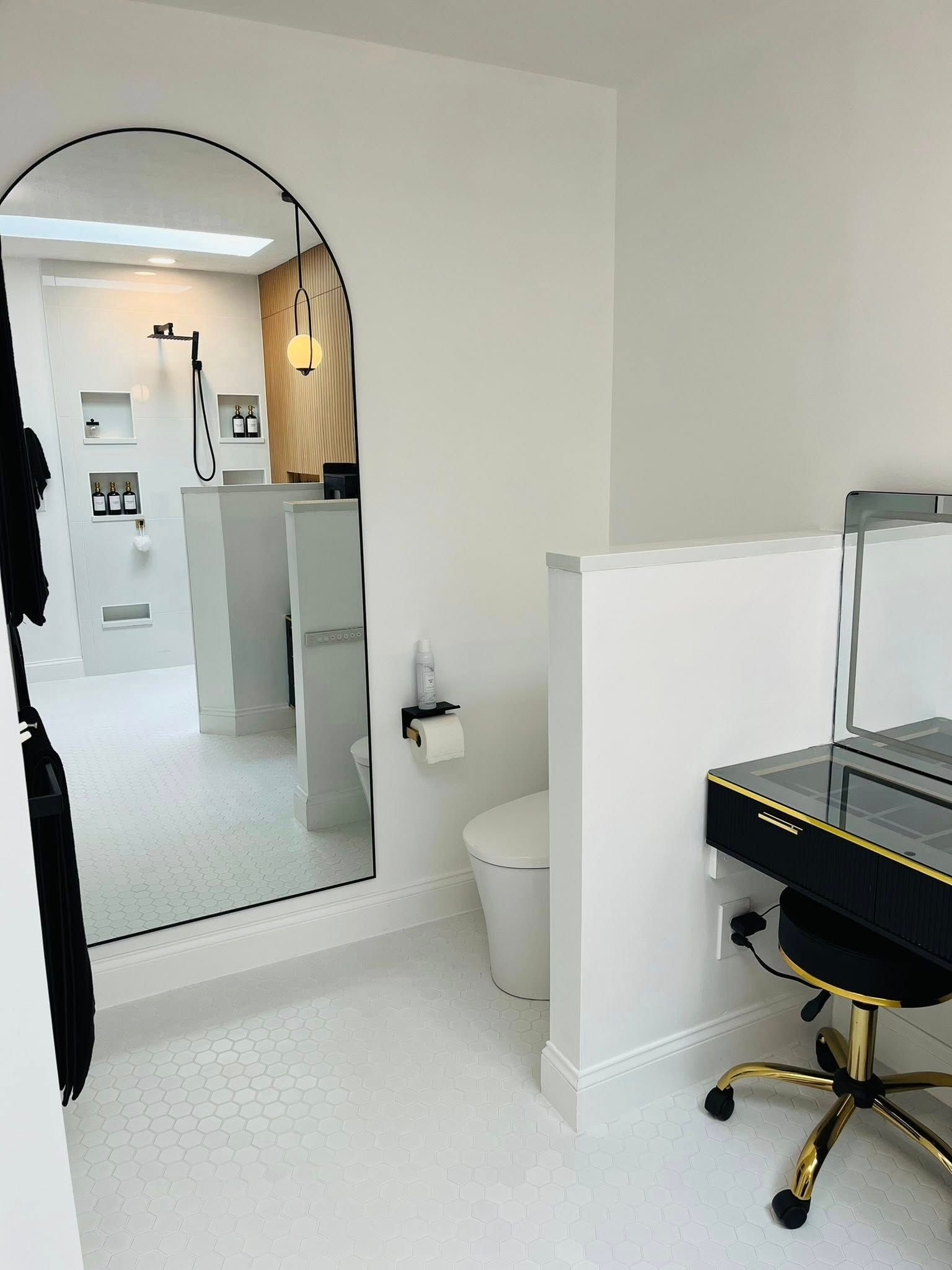 Small white room with arched mirror, sink, toilet, and black desk with gold chair