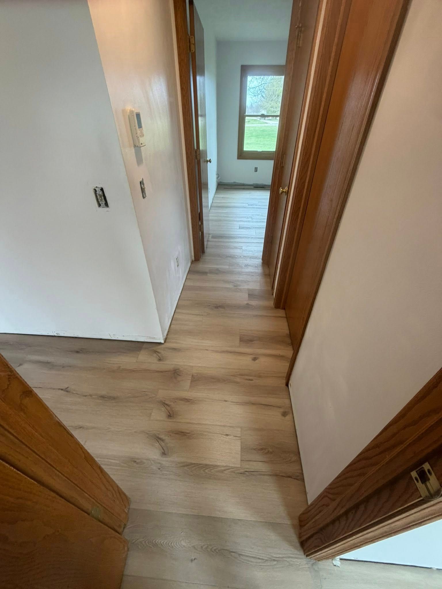 Narrow hallway with wood flooring and white walls, leading to a window at the far end.