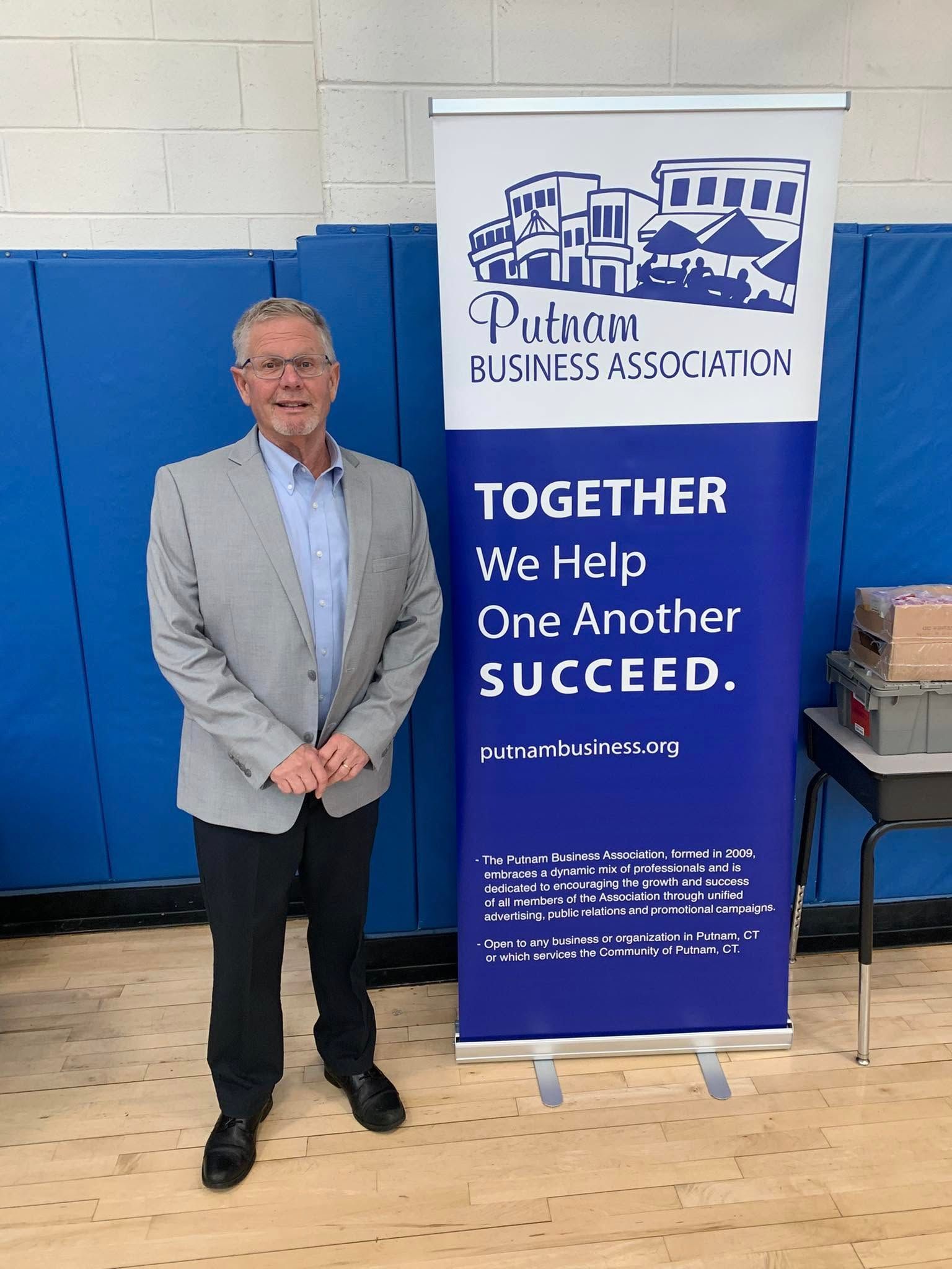 Man in a suit stands beside a banner with the Public Business Association logo and the phrase