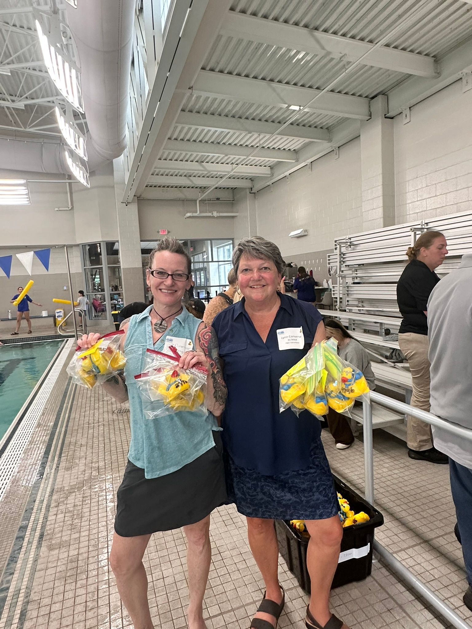 Two women smiling, holding bags of rubber ducks, next to a swimming pool.