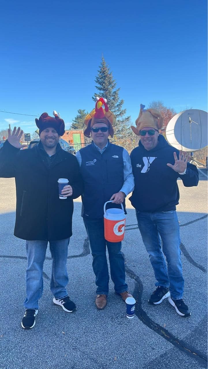 Three men wearing turkey hats, waving and smiling outside on a sunny day, two holding drinks.