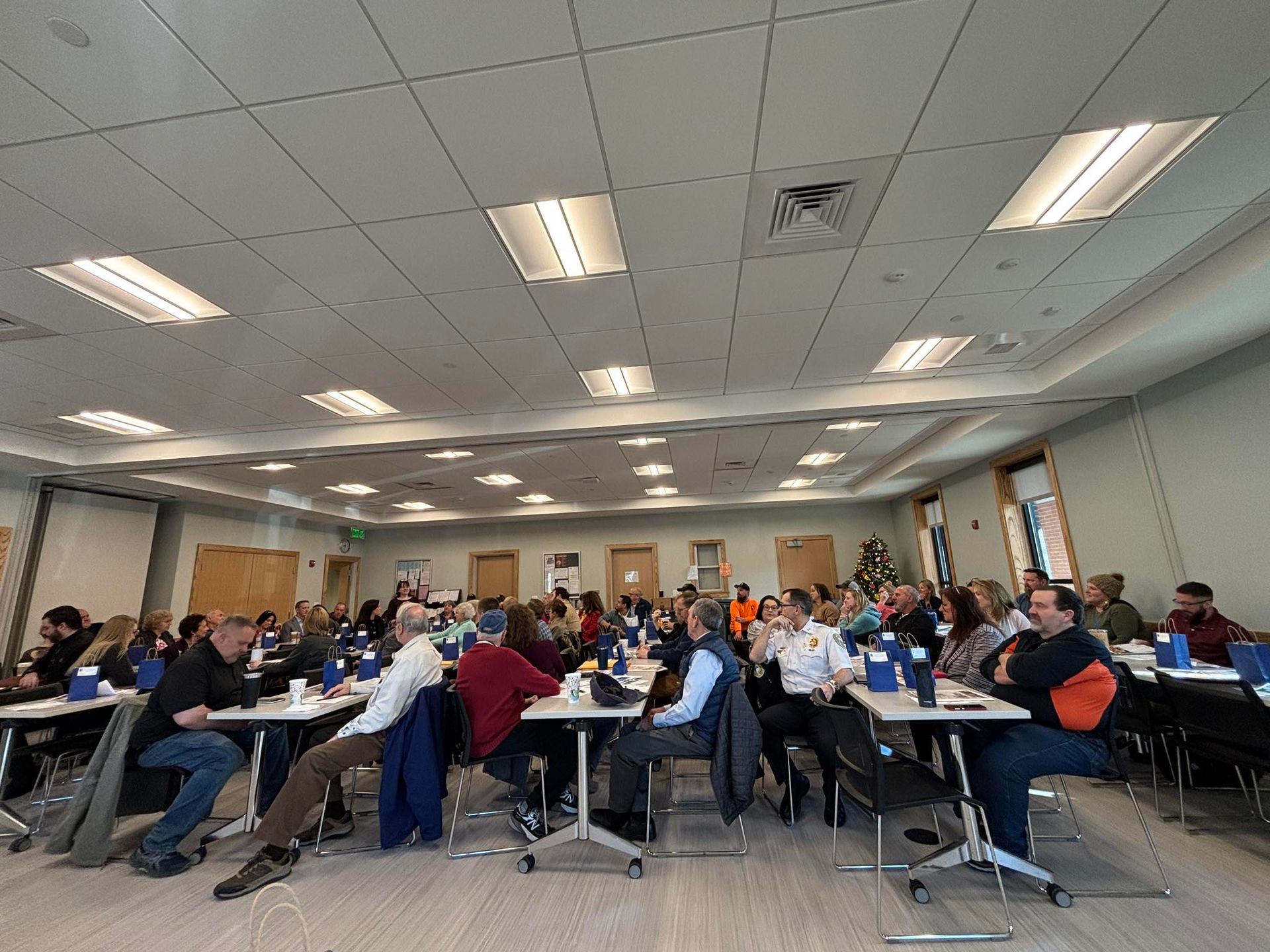 Large room with people seated at tables, likely in a meeting or conference. Overhead lighting.