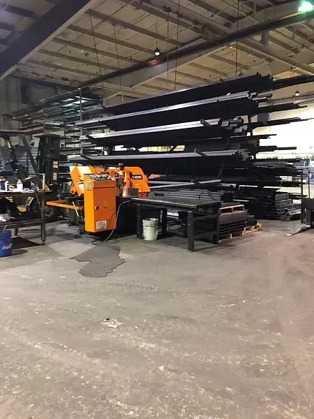 Metal beams stacked in a warehouse with machinery and an orange cutting machine.