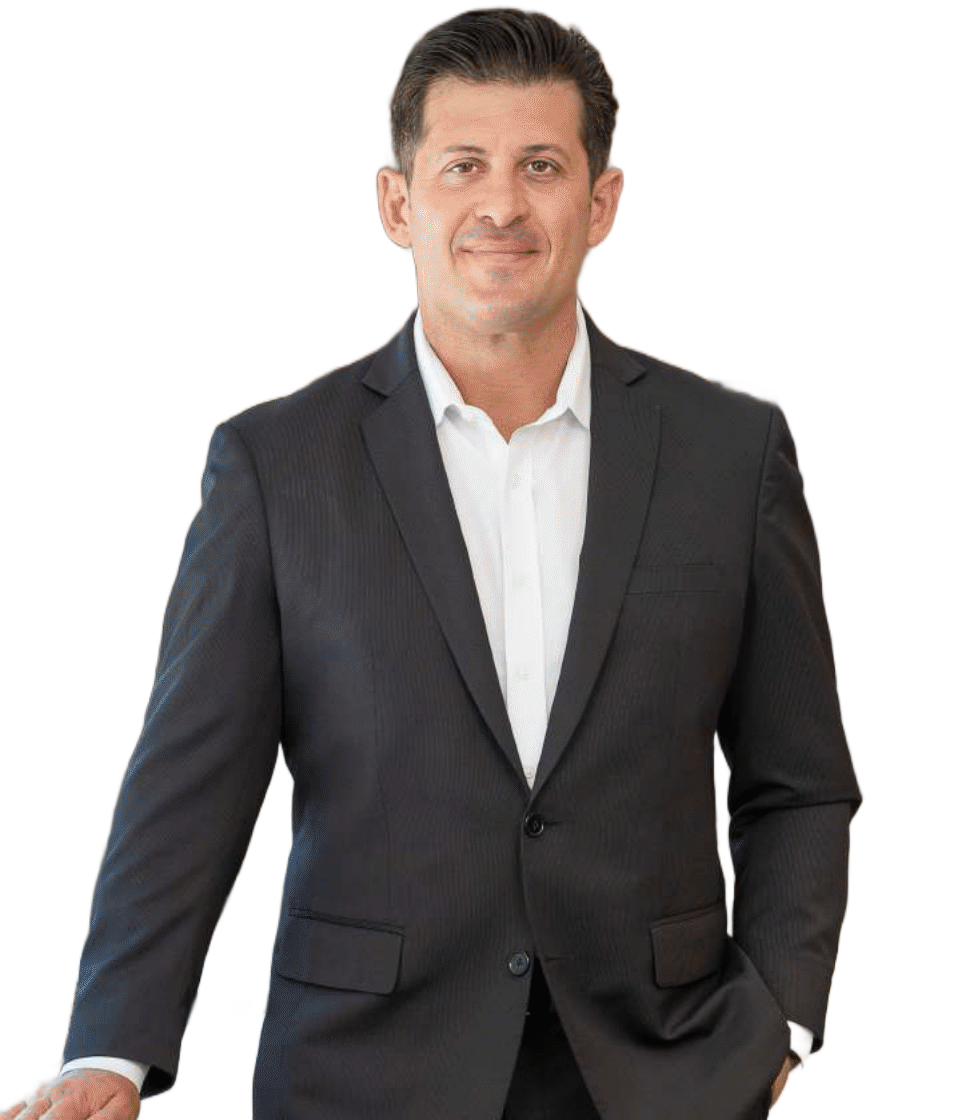 Man in a suit, leaning on a surface, smiling, white shirt, dark jacket, indoor setting.