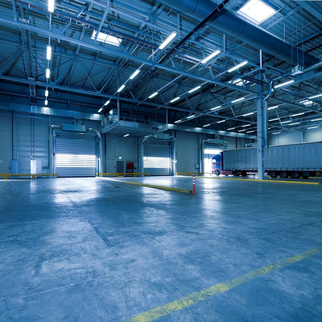Large, blue-toned warehouse interior. Open loading doors, truck parked in the distance, bright ceiling lights.