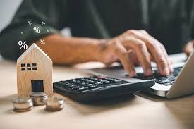Person working on laptop, calculating mortgage with calculator and house figurine, coins.