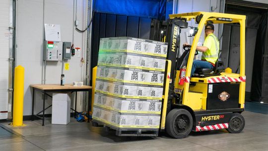 A man is driving a forklift in a warehouse carrying a stack of boxes.