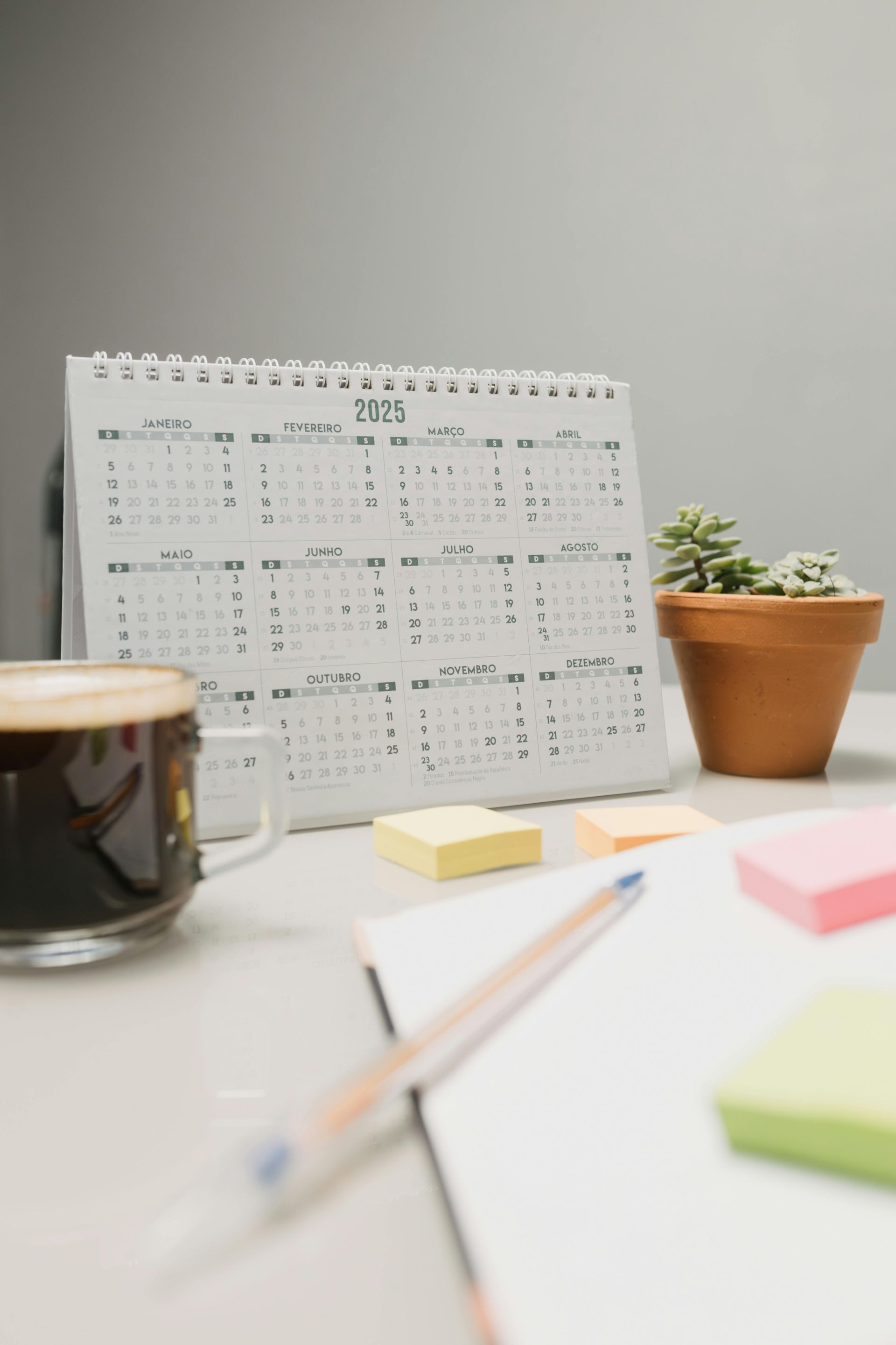 A calendar , sticky notes , a cup of coffee and a potted plant are on a desk.