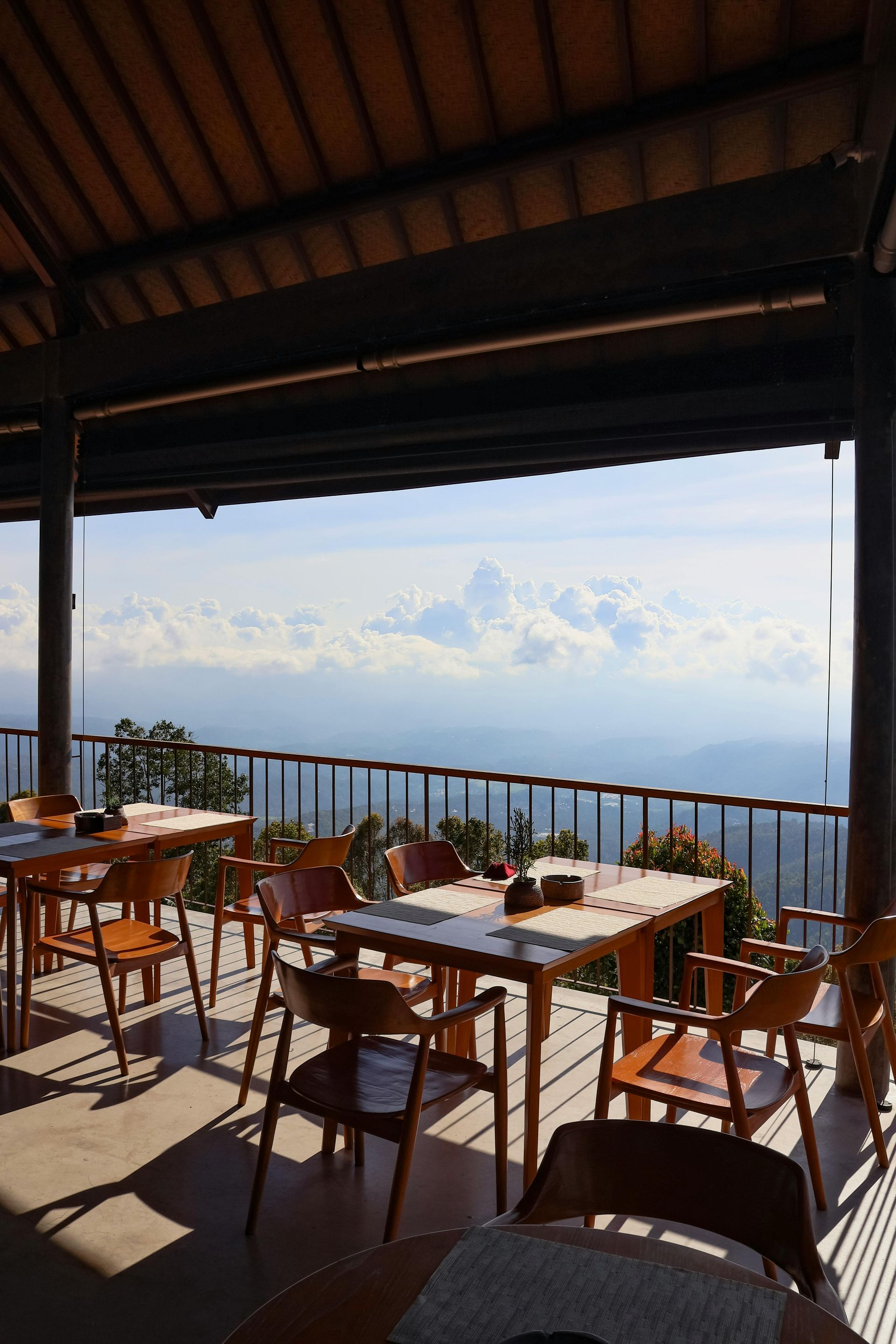 Wooden tables and chairs on a porch overlooking a cloudy, mountainous landscape under a wood-beamed ceiling.