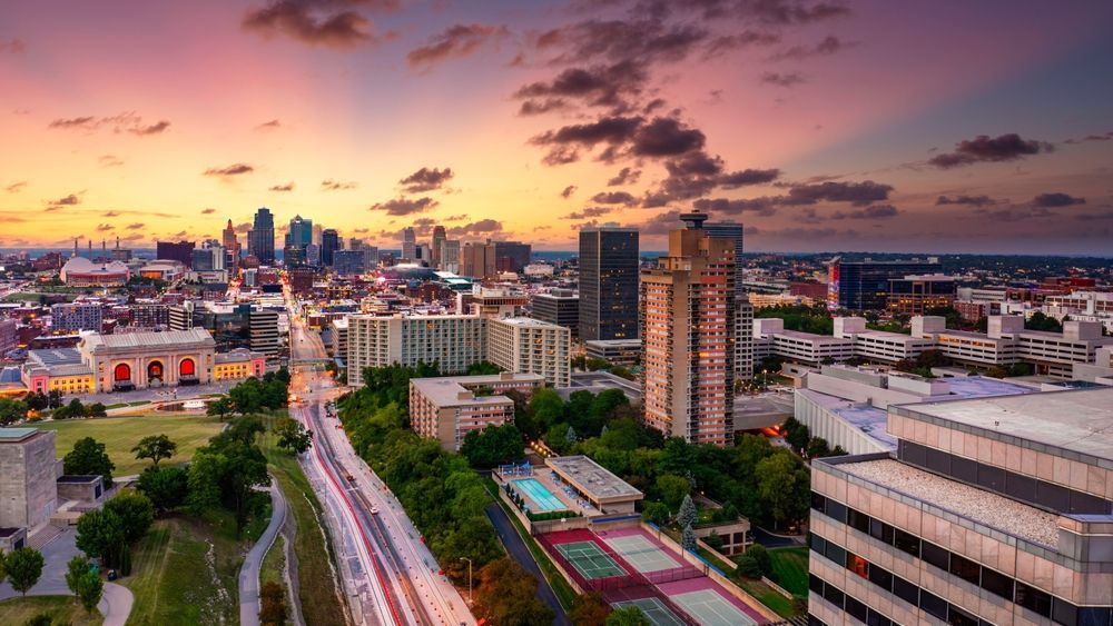 Kansas City skyline at sunset, orange and purple sky, city lights, and tall buildings.