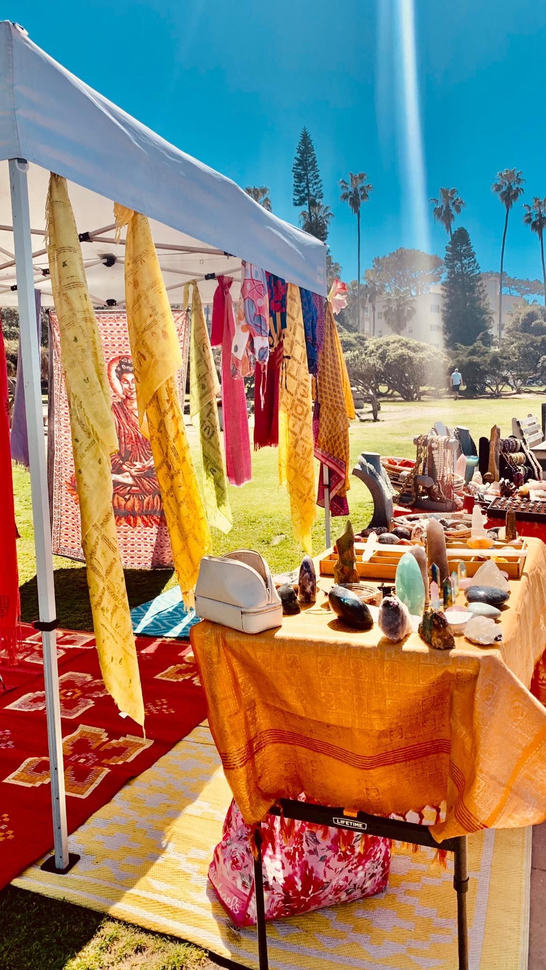 Outdoor market with colorful textiles, jewelry, and crystals on display under a white tent, bright sunny day.