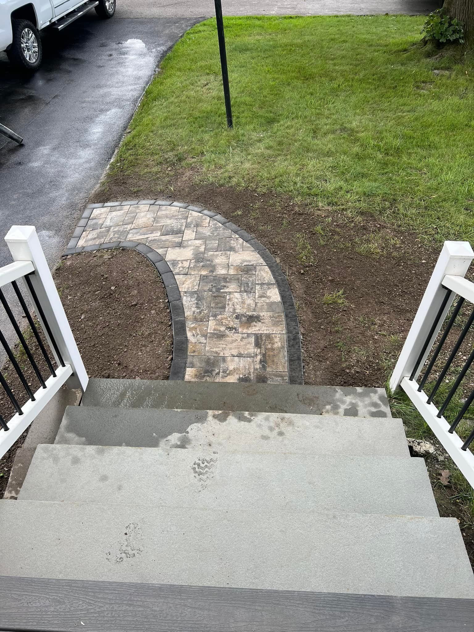 Concrete steps leading to a brick pathway. The pathway curves towards a green lawn. White railings flank the steps.