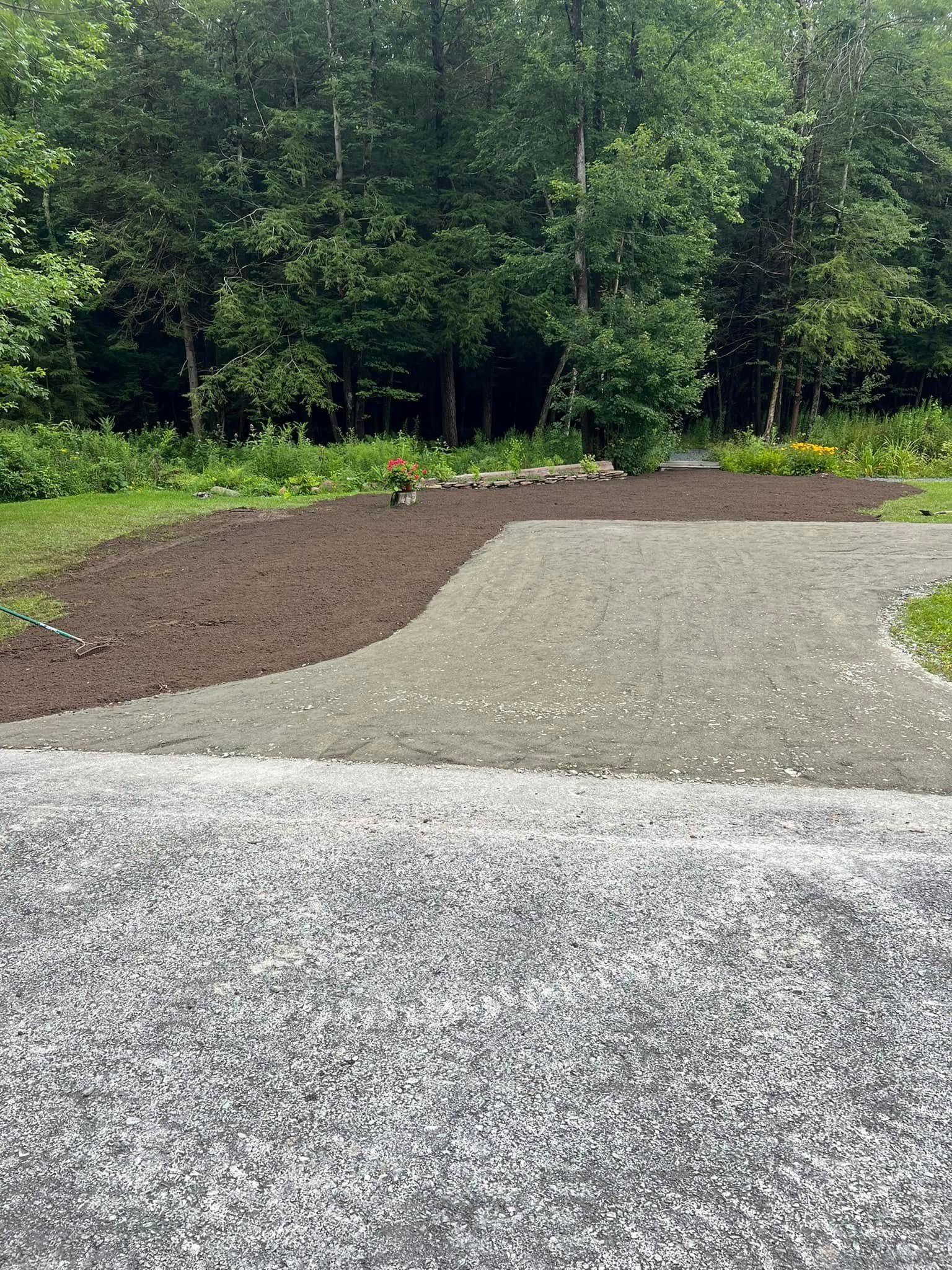 Gravel driveway transitions to a landscaped area with dirt and lighter gravel, leading to a wooded backdrop.