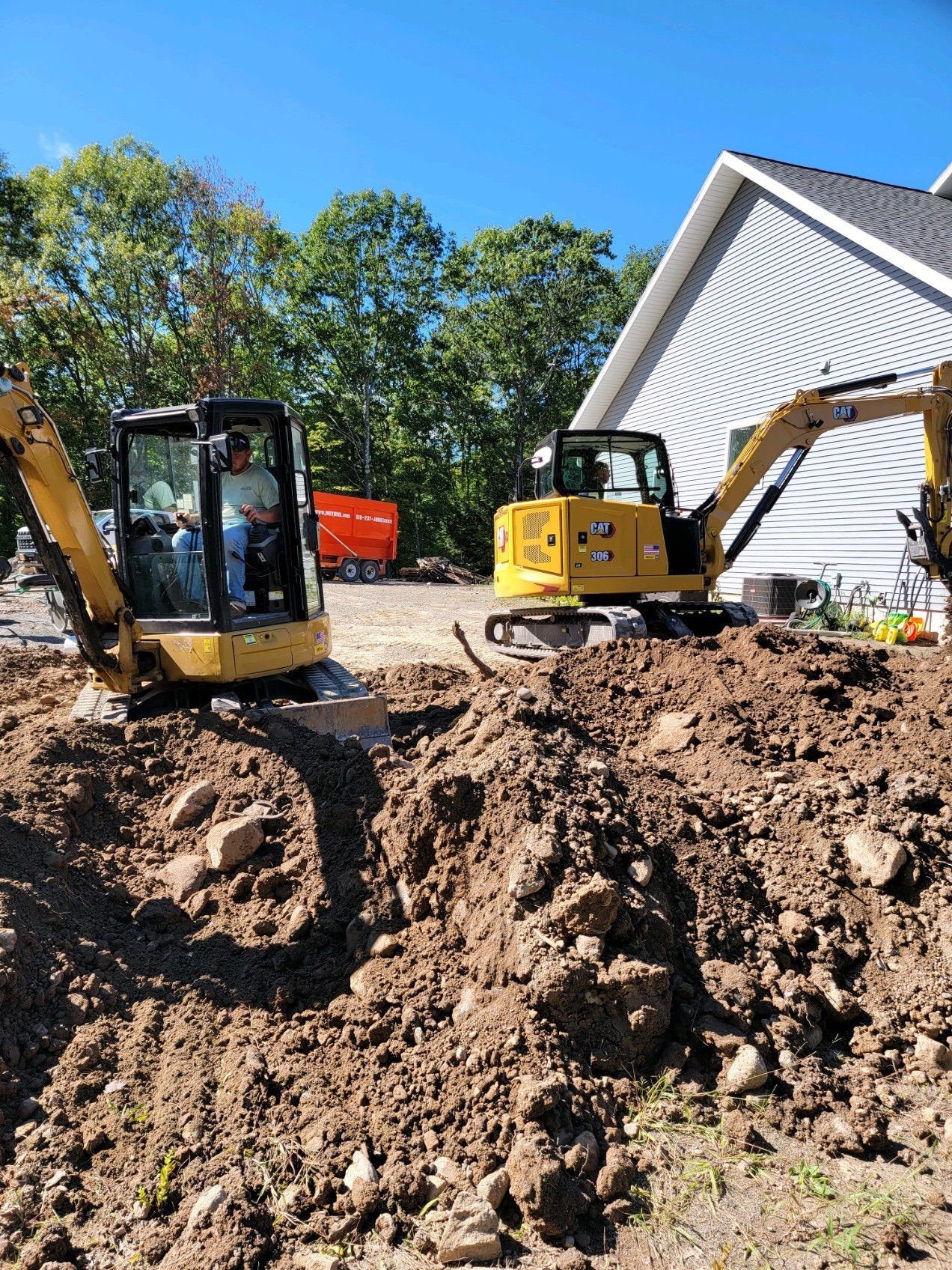 Construction site with an excavator digging near apartment buildings. Gravel driveway and a grassy hill.