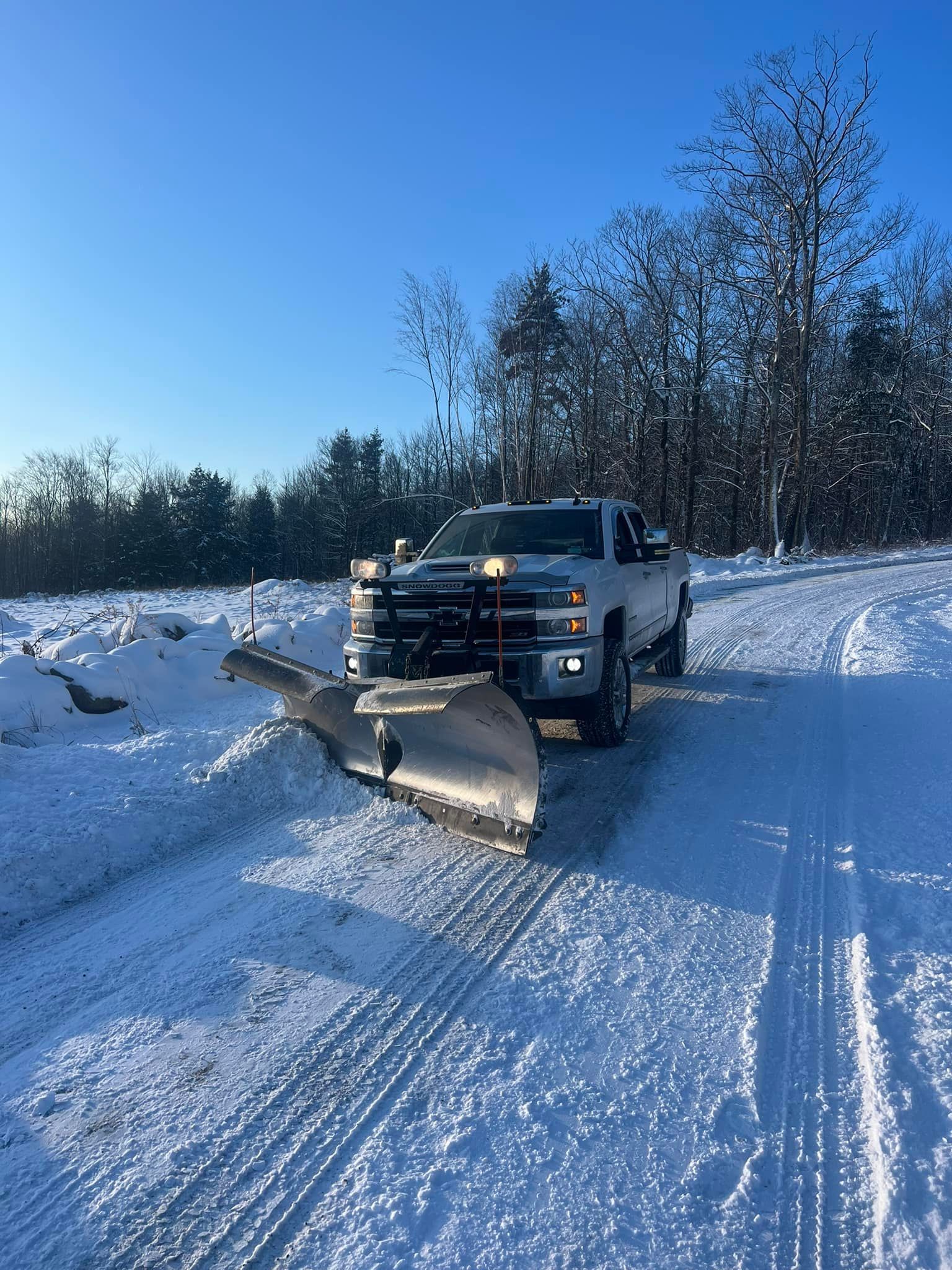 Yellow snowplow clearing a snow-covered parking lot with cars. Brick building and trees in the background, overcast day.