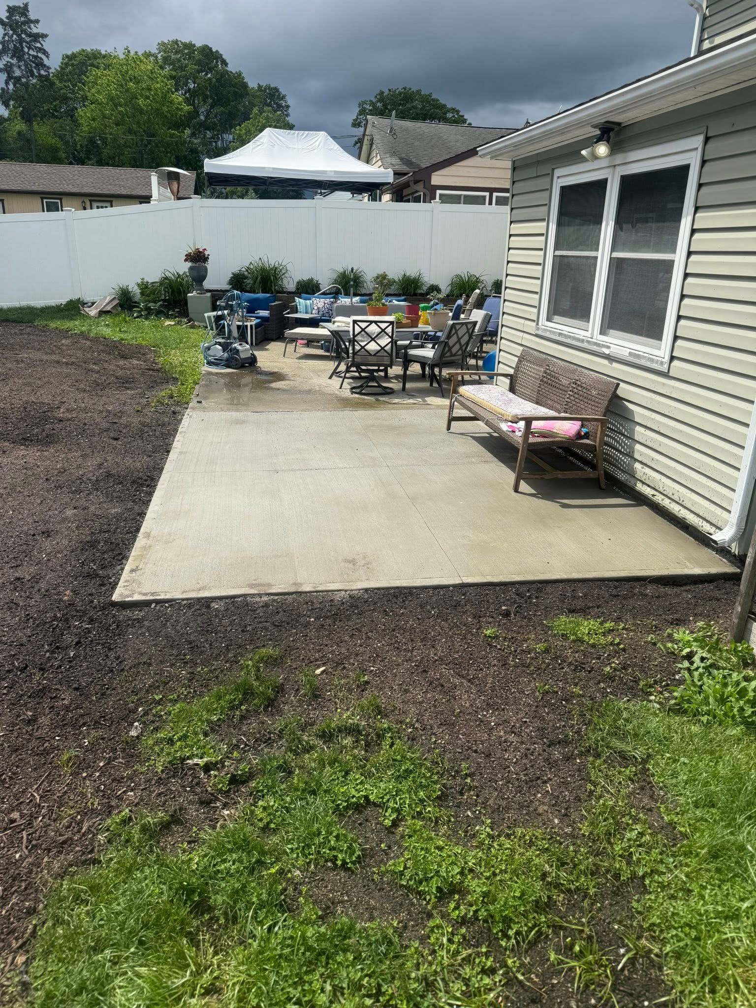 Backyard with concrete patio, furniture, and mulch. House siding and white fence in the background. Overcast sky.