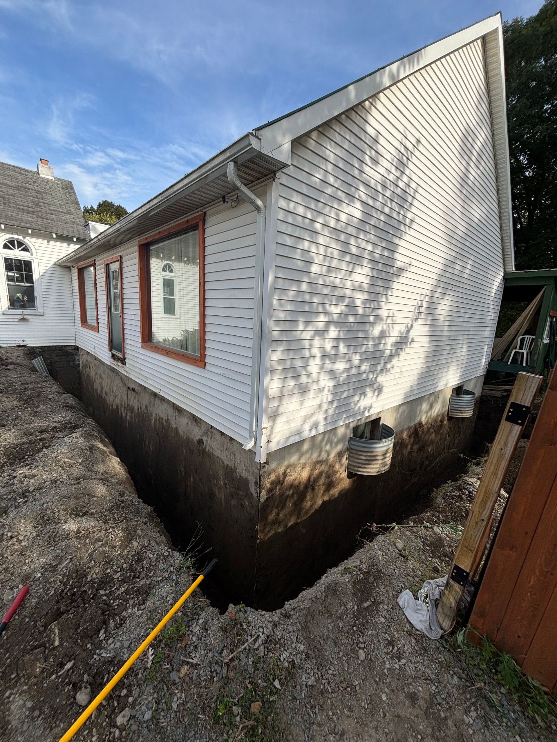 A house with white siding and a trench dug around its foundation; daytime.